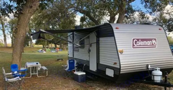 Enjoy an RV rental! This Coleman travel trailer is set up at a leafy campsite with an extended awning, chairs, & picnic table.. Coleman Lantern 2023