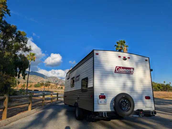 This Coleman travel trailer rental is parked with mountains & palm trees in the background, showing its rear & spare tire.. Dutchmen Coleman Lantern LT 2023