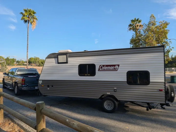 A striped Coleman travel trailer (RV rental) is towed by a blue pickup truck, parked on asphalt under palm trees.. Dutchmen Coleman Lantern LT 2023