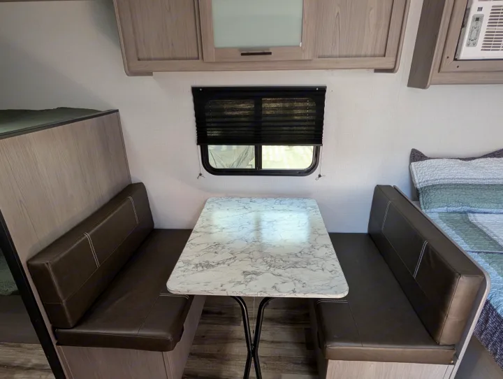 RV rental interior: Bright dinette with marble-patterned table, dark benches, and adjacent bunk/bed areas, perfect for travel.. Dutchmen Coleman Lantern LT 2023