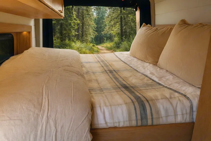 Cozy RV rental bed with a plaid blanket, looking out to a serene forest path. Perfect for nature travel.. MERCEDES-BENZ Sprinter 2500 AWD 2024