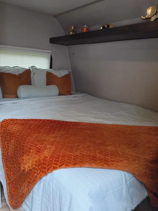 Cozy RV rental bedroom featuring white bedding, an orange throw, pillows, and a decorative shelf above the bed.. Keystone RV Springdale 2019