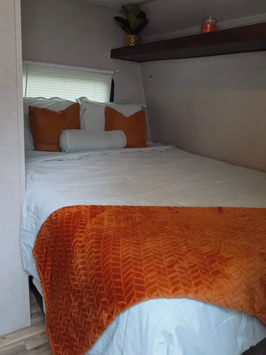 Cozy RV rental bedroom featuring a neatly made bed with light blue sheets, orange pillows and blanket, plus a shelf with decor.. Keystone RV Springdale 2019
