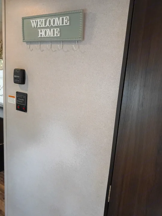 RV rental interior: "Welcome Home" key holder, Suburban thermostat, and Command Center panel. Dark wood door and textured wall.. Keystone RV Springdale 2019