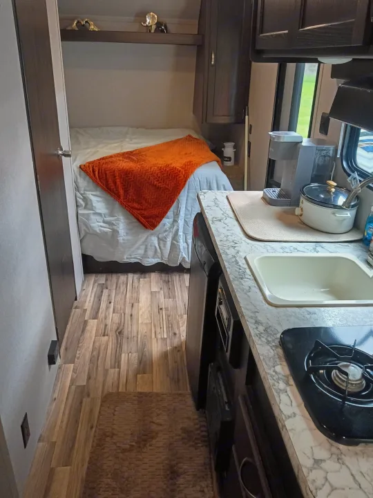 This inviting rv rental features a compact kitchen with sink, stove, coffee maker, and a cozy bed with an orange blanket.. Keystone RV Springdale 2019