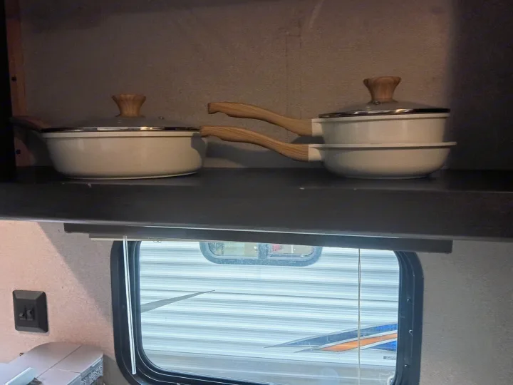 Stacked pots with wooden handles and glass lids on a shelf in an RV rental kitchen, with a window showing the exterior.. Keystone RV Springdale 2019