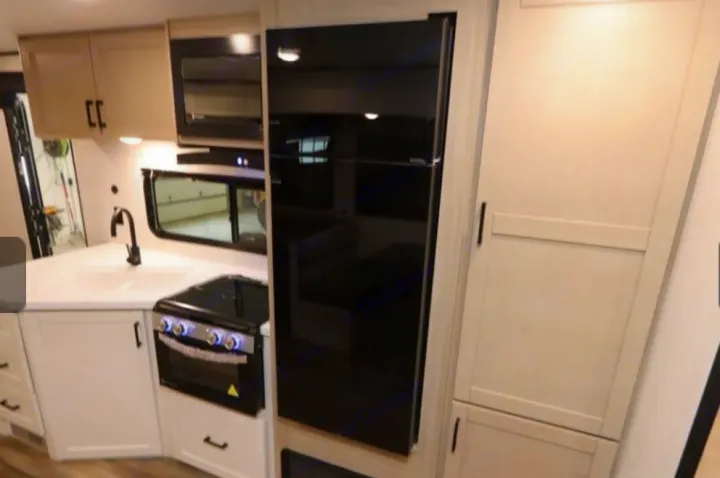 Modern rv rental kitchen showing a corner sink, stove, microwave, sleek black refrigerator, and light-colored pantry storage.. Highland Ridge RV Colt 2026