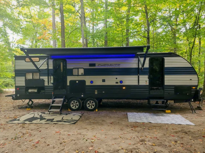 Spacious Cherokee rv rental set up at a wooded campsite, featuring an extended awning with blue lights, stairs, and ground mats.. Forest River Cherokee 2021