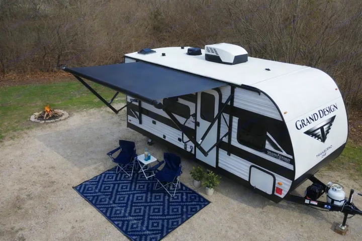 Overhead view of a Grand Design travel trailer (rv rental) with an extended awning, outdoor rug, chairs, and a fire pit.. Transcend One 151BH 2026