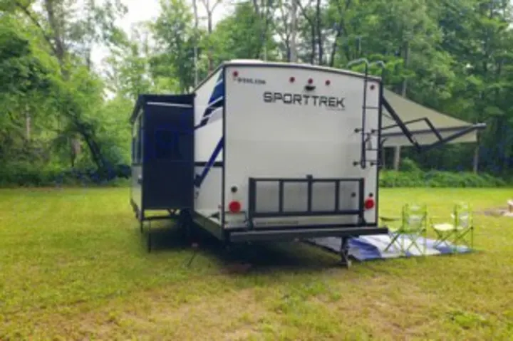 This RV rental, a white SportTrek travel trailer, is set up for camping with an awning & slide-out extended in a grassy, wooded area.. Venture SportTrek 2021
