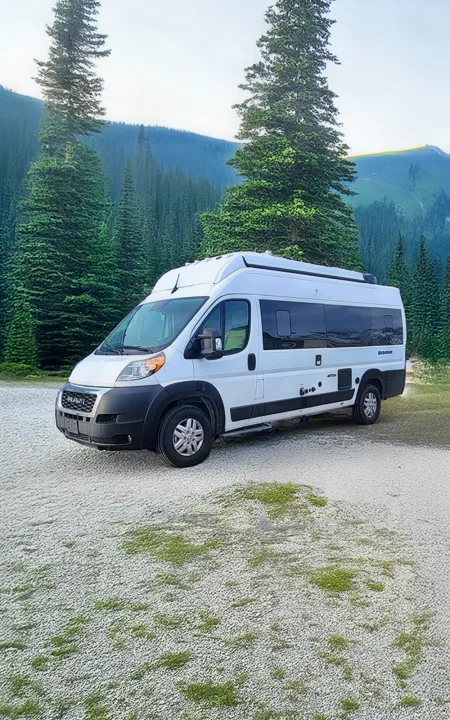 White RV rental camper van parked on gravel, surrounded by pine trees and mountains. Ideal for nature adventures.. Thor Motor Coach Sequence 2023