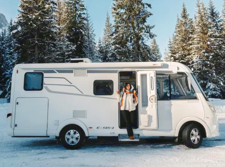 Exploring snowy landscapes in an RV rental. A woman in a scarf stands at the entrance of her white campervan amidst snow-covered trees.. HYMER EXISIS I 474 2025