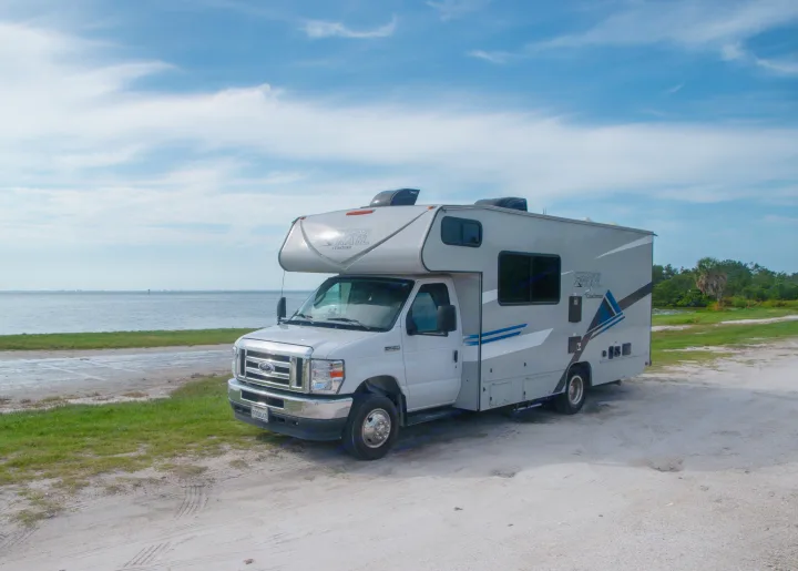 This grey rv rental motorhome is parked on a sandy area beside calm water, with a blue sky above. Perfect for travel adventures.. Coachmen Cross Trail 2023