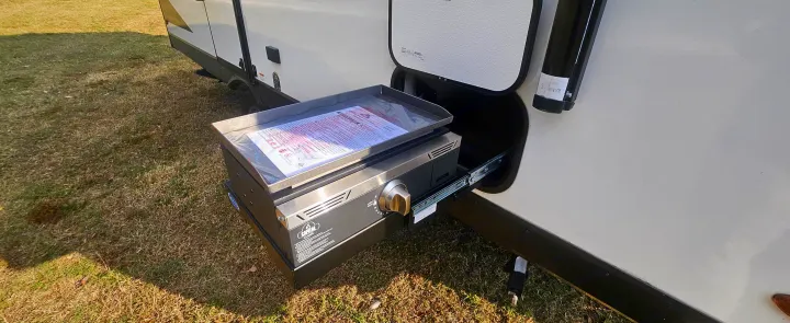 RV rental with a pull-out outdoor griddle for convenient camping meals. Manual rests on the cooktop, parked on grass.. Grand Design Imagine AIM 2023