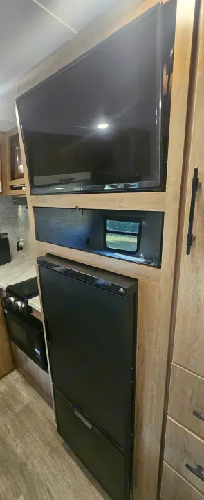 Modern RV rental interior: TV over a compact fridge, adjacent to a stove. Efficient kitchen setup for travel.. Grand Design Imagine AIM 2023