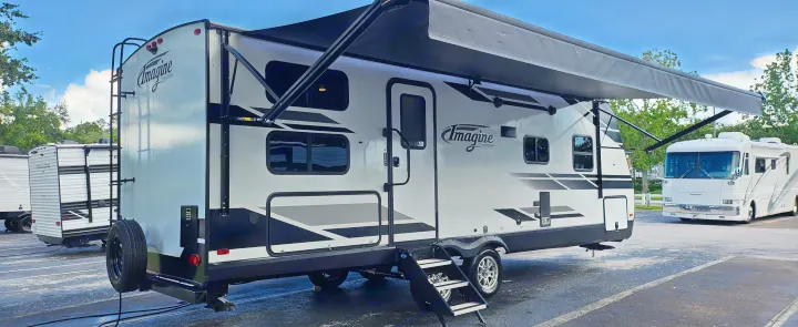 White travel trailer RV rental with extended awning & steps, parked with other recreational vehicles in an outdoor lot.. Grand Design Imagine 2021