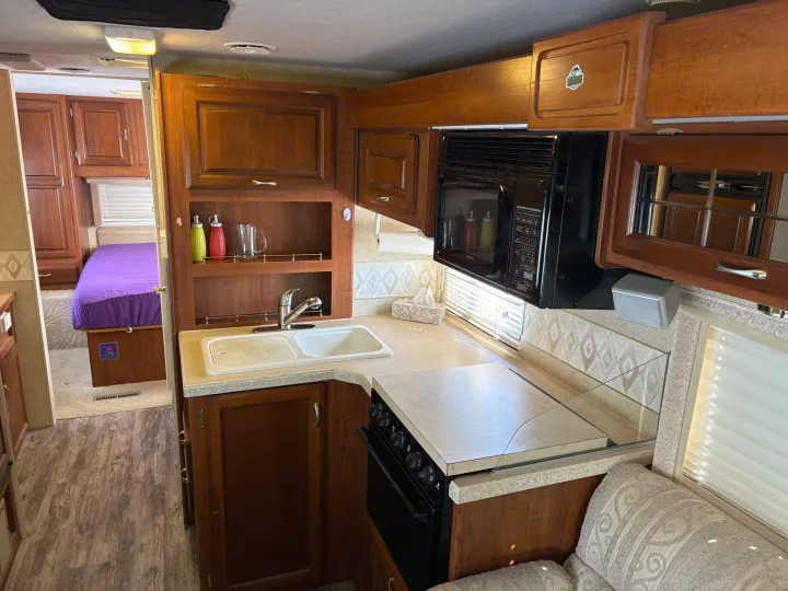 View inside an RV rental showing a full kitchen with microwave, stove, sink, and wood cabinets, plus a peek into the bedroom with a purple bed.. Jayco Granite Ridge 2004
