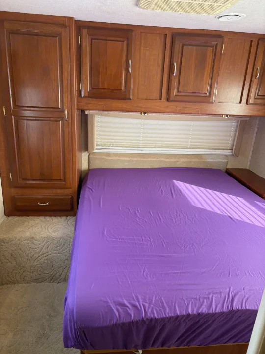 RV rental bedroom with a bed in bright purple sheets, overhead wooden cabinets, and a tall wardrobe. Sunlight falls across the bed.. Jayco Granite Ridge 2004