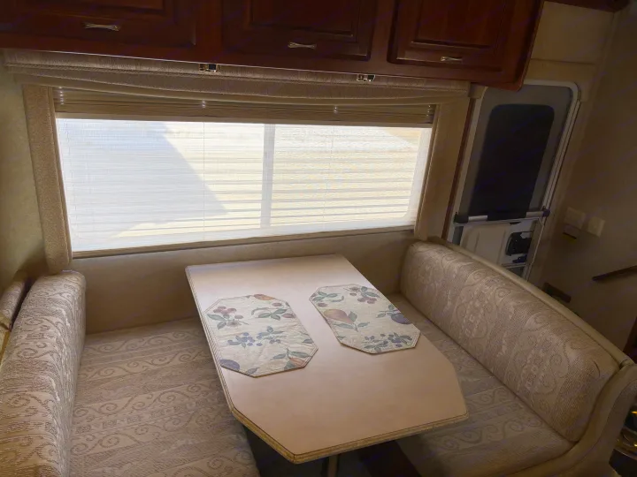 Cozy RV rental dinette featuring patterned benches, a light-colored table, window with blinds, and overhead cabinets.. Jayco Granite Ridge 2004