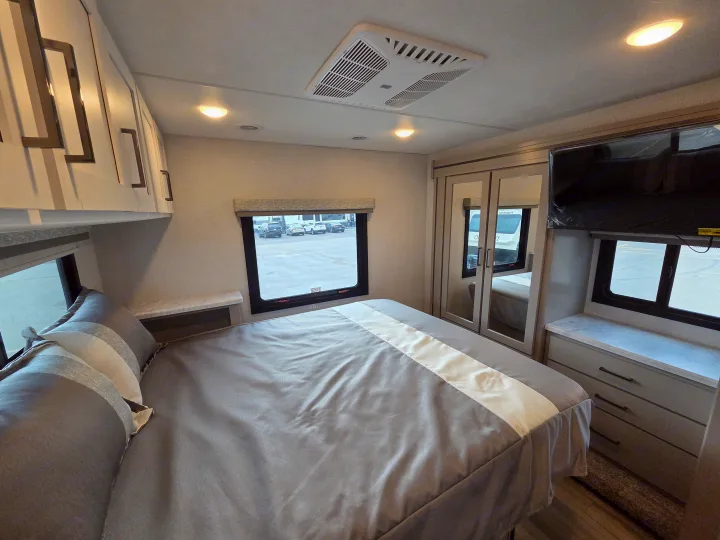 This RV rental bedroom features a comfy bed with gray and white bedding, overhead cabinets, a window, TV, and mirrored closet.. Four Winds 31E 2026