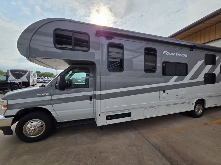 Gray and white Four Winds Class C RV rental, side view. Parked outdoors under a cloudy sky, with another RV in the background.. Four Winds 31E 2026
