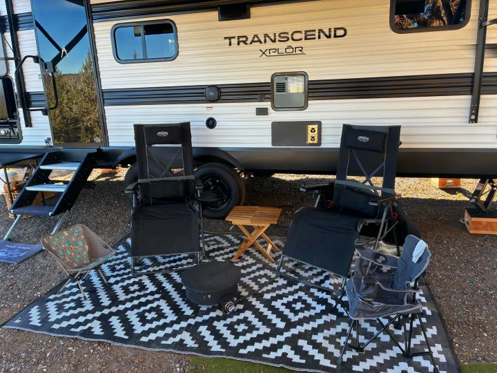 Cozy RV rental outdoor living area with chairs, fire pit, and patterned rug at a scenic campsite.. Grand Design Transcend Xplor 2026