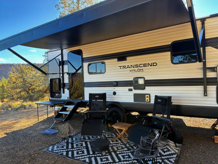 Enjoying the outdoors! This rv rental is set up with an extended awning, chairs, and a portable fire pit at a picturesque campsite.. Grand Design Transcend Xplor 2026
