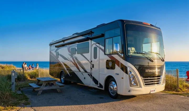 Scenic rv rental parked at a coastal campsite. A modern motorhome, picnic table, and people by the ocean.. Thor Motor Coach Windsport 2022