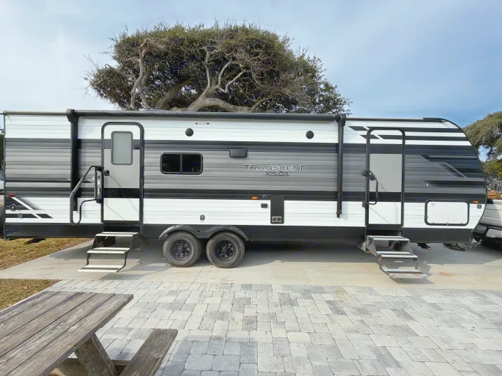 A large grey, black, and white Transcend Xplor RV rental is parked on a site with steps, pavers, and a picnic table under a cloudy sky.. Grand Design Transcend 265BH 2022
