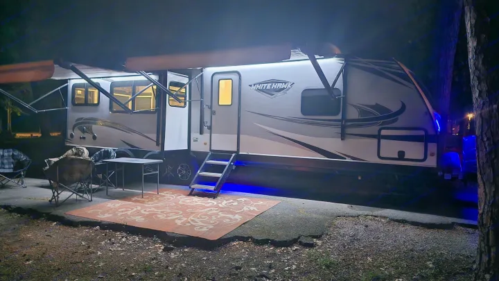 Cozy night view of a brightly lit rv rental travel trailer at a campsite with awning and blue underglow lights, outdoor rug & chairs.. Jayco White Hawk 2020