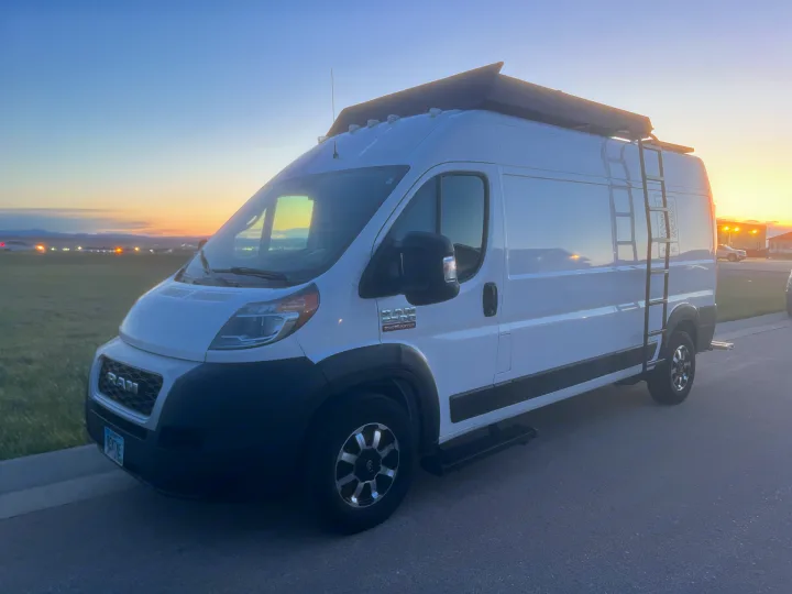 White Ram ProMaster RV rental van, customized with a ladder, awning, and side steps, parked at sunset.. Ram Promaster 2500 2019