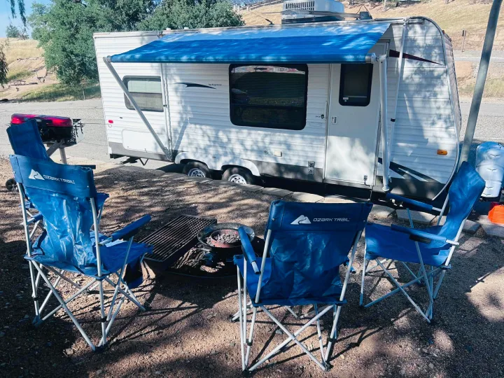 Sunny RV rental campsite: a white travel trailer under an awning, with blue Ozark Trail chairs and a fire pit. Ready for camping!. Keystone RV Hideout 2009