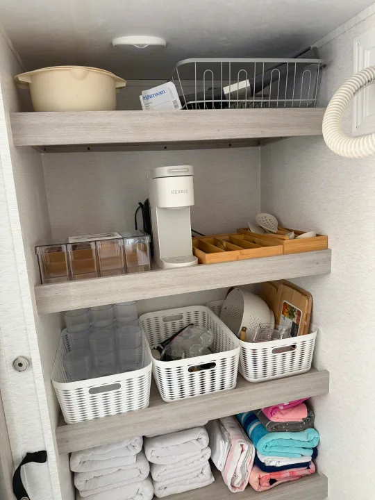 RV rental pantry shelves displaying a Keurig, organized kitchenware (utensils, glasses, containers), and neatly folded towels.. Crossroads RV Sunset Trail Sport 2025