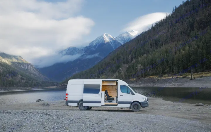 RV rental adventure! A white camper van with an open side door on a rocky lake shore, backed by majestic mountains.. Sprinter Mercedes 2018