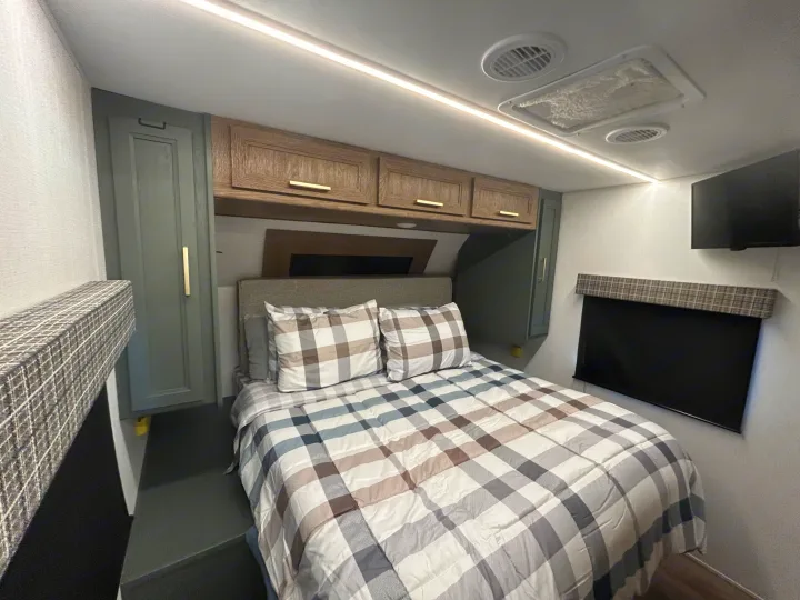 RV rental bedroom: plaid bedding, modern overhead cabinets, wardrobe, wall-mounted TV, and bright lighting.. Heartland RVs Eddie Bauer 2024
