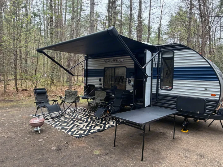 An RV rental provides a cozy campsite setup with an extended awning, several chairs, a grill, and a table amidst a forest.. Forest River Cherokee Wolf Pup 2021
