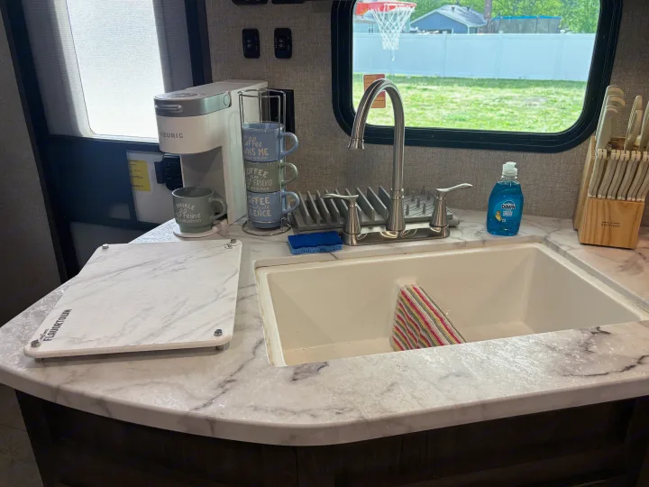 RV rental kitchen: Keurig, sink, mugs, and a cutting board on a marble counter. Outside, a yard with a basketball hoop is seen.. Jayco Jay Flight 2020