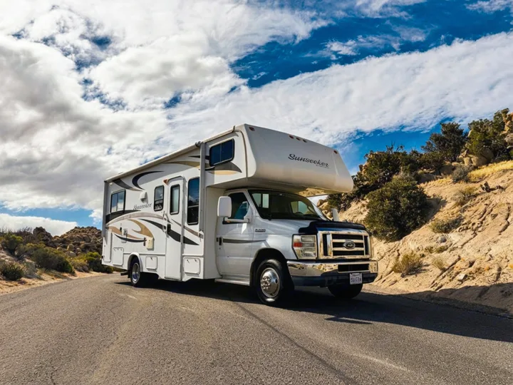 A white Sunseeker RV rental is parked on a winding road amidst rocky desert hills under a dramatic cloudy sky.. Forest River Sunseeker 2012