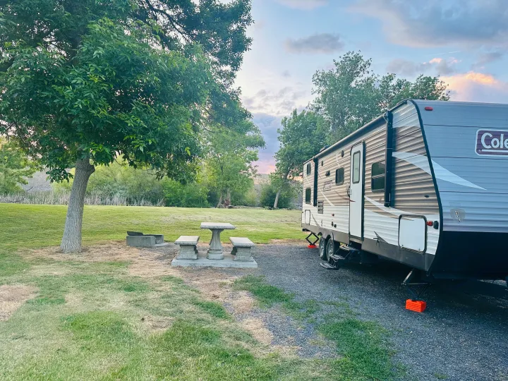 RV rental parked at a serene campsite with a stone picnic table, fire pit, and lush trees, perfect for outdoor relaxation.. Dutchmen Coleman Lantern 2019