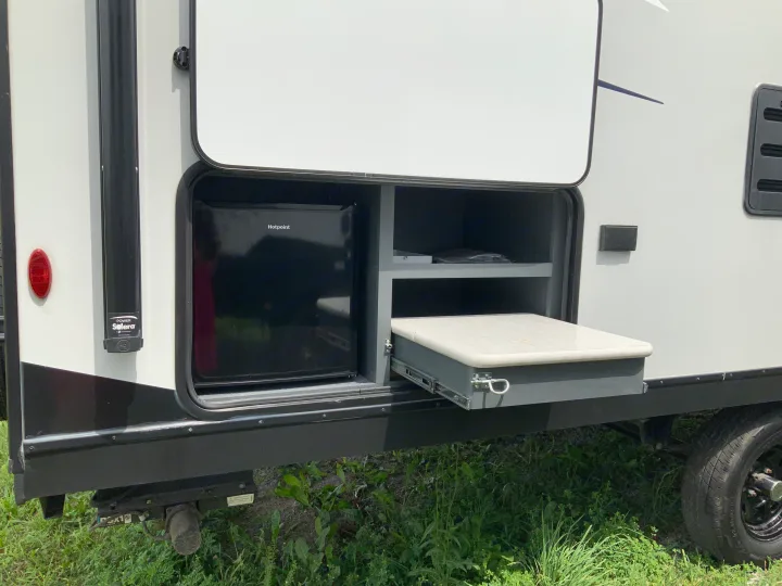 An RV rental's exterior compartment reveals a Hotpoint mini-fridge, shelves, and an extended pull-out counter.. Keystone RV Bullet Crossfire 2022