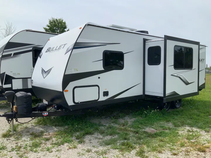 White Bullet Crossfire travel trailer, an RV rental with its slide-out extended, parked on gravel with some green grass.. Keystone RV Bullet Crossfire 2022