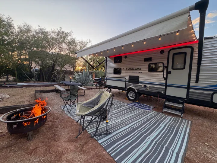 RV rental campsite at dusk, featuring a travel trailer with illuminated awning, a crackling fire pit, and camping chairs.. Coachmen 184BHS 2021