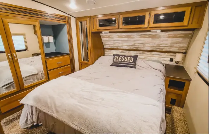Enjoy this RV rental's comfortable bedroom with a "BLESSED" pillow, plank-style headboard, mirrored wardrobe, and overhead storage.. Coachmen Chaparral 2018