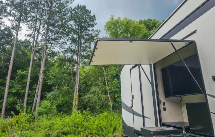 RV rental featuring an outdoor kitchen and entertainment area with a TV, surrounded by tall pine trees and lush greenery.. Coachmen Chaparral 2018
