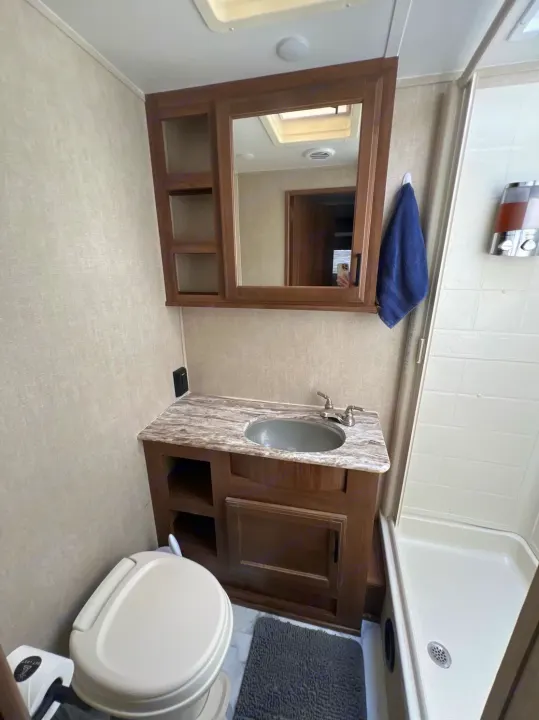 An RV rental bathroom, showing a toilet, vanity with sink, mirrored cabinet, and shower stall with a blue towel.. Gulf Stream Conquest 2019