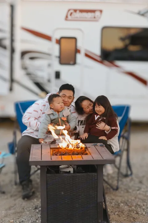 A family of four sits around a fire pit by their Coleman RV rental, enjoying a cozy outdoor evening together.. Thor Motor Coach Coleman 2023