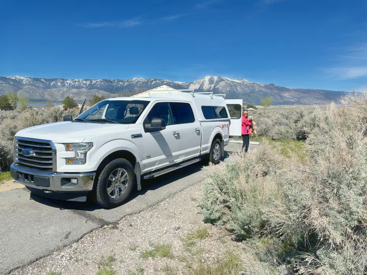 An RV rental journey: White truck with camper, a person holding a dog, scenic snowy mountains, and a lake.. Ford Ford F150 2017
