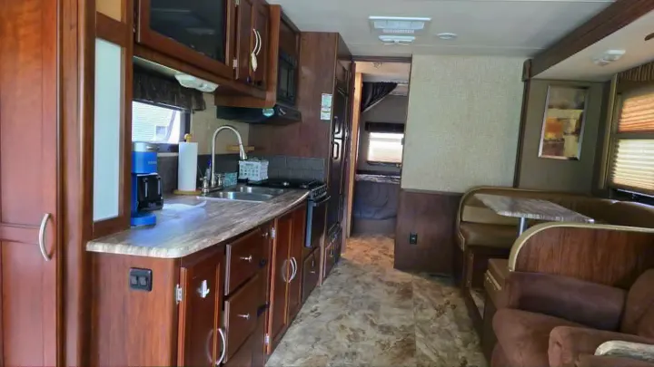Spacious rv rental interior showing a kitchen, dining booth, and living area with a brown armchair, featuring wood cabinetry and light flooring.. 1F65F5DY3E0A15748 Pursuit 31 BD 2015