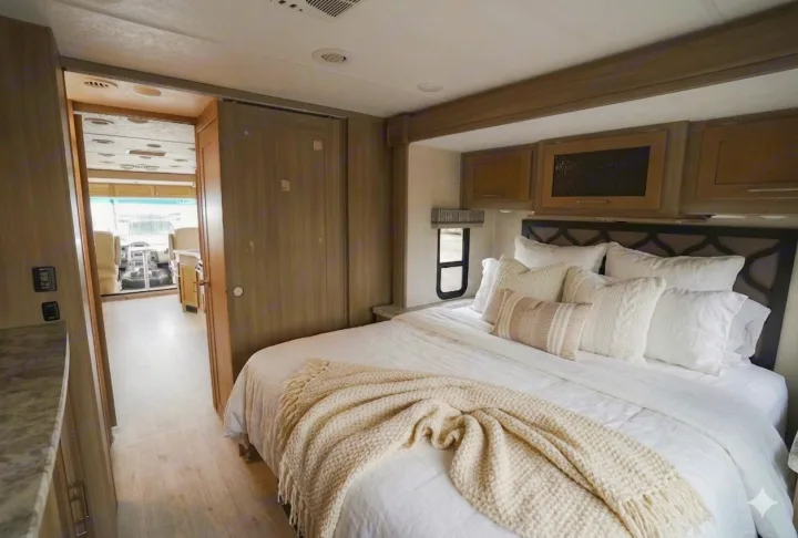 Spacious RV rental bedroom with modern decor, white bedding, and a view into the cockpit area.. Coachmen Pursuit 2019