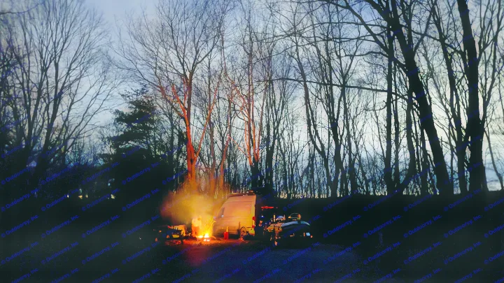 Dusk at an **RV rental** campsite: a bright campfire illuminates a person & camper van, silhouetted by bare trees.. Ford Custom Transit 2020